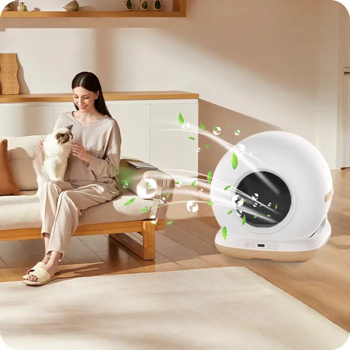 A woman sitting on a couch petting a fluffy cat next to a white automatic litter box emitting green freshness graphics.