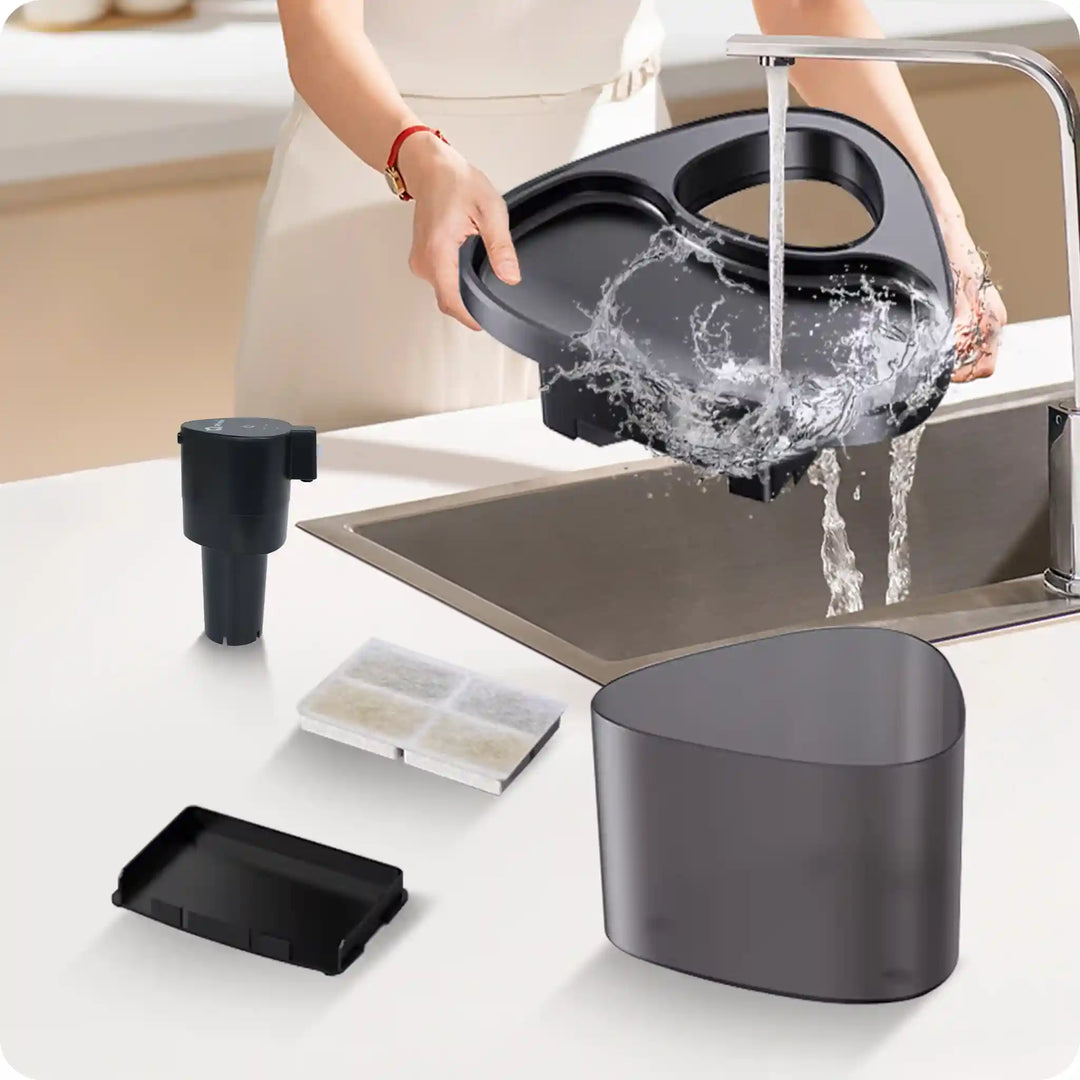Person rinsing pet water fountain parts at sink with filter and black base pieces spread out.