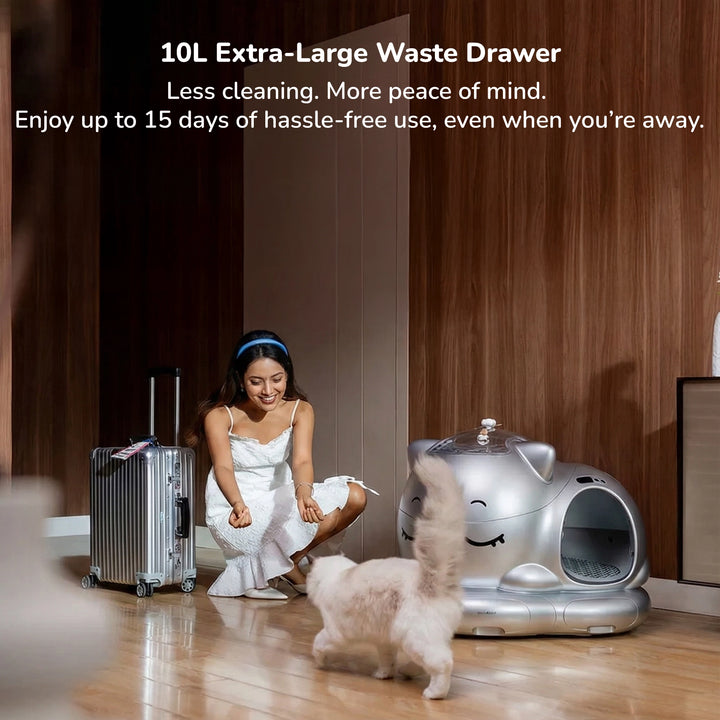 Woman crouching near a silver smart cat litter box with an extra‑large waste drawer and a fluffy cat in a wood‑paneled room.